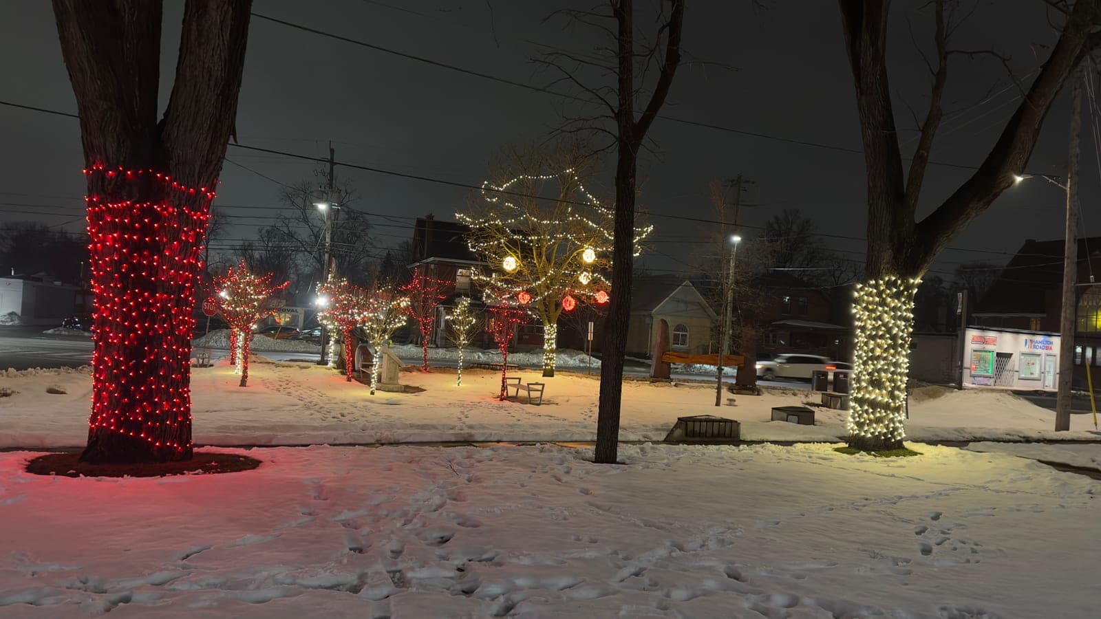 Downtown park with trees wrapped in red and warm-white Christmas lights and glowing red ornament balls over snow