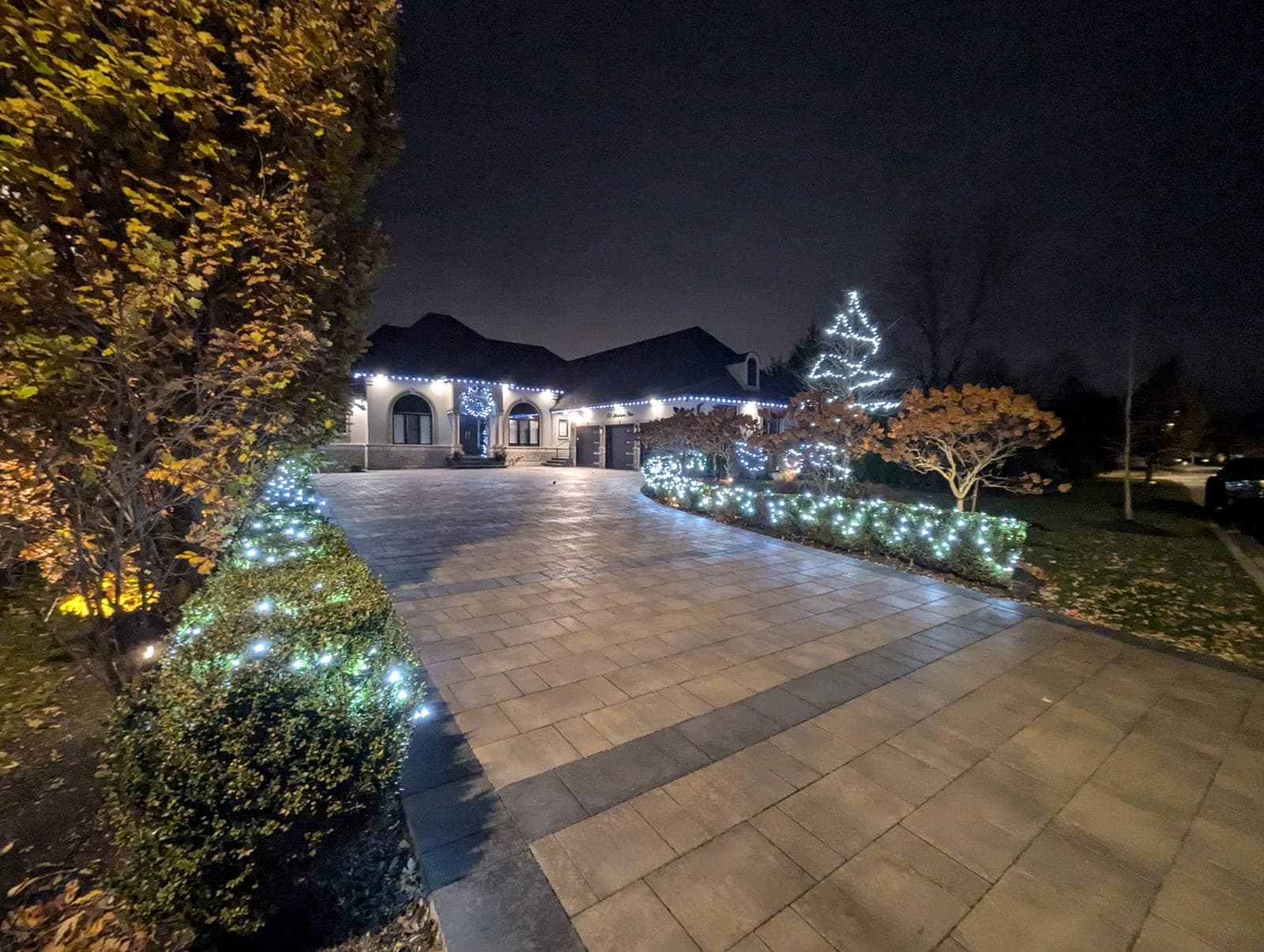Estate driveway lined with cool-white lit shrubs leading to a home with roofline Christmas lights