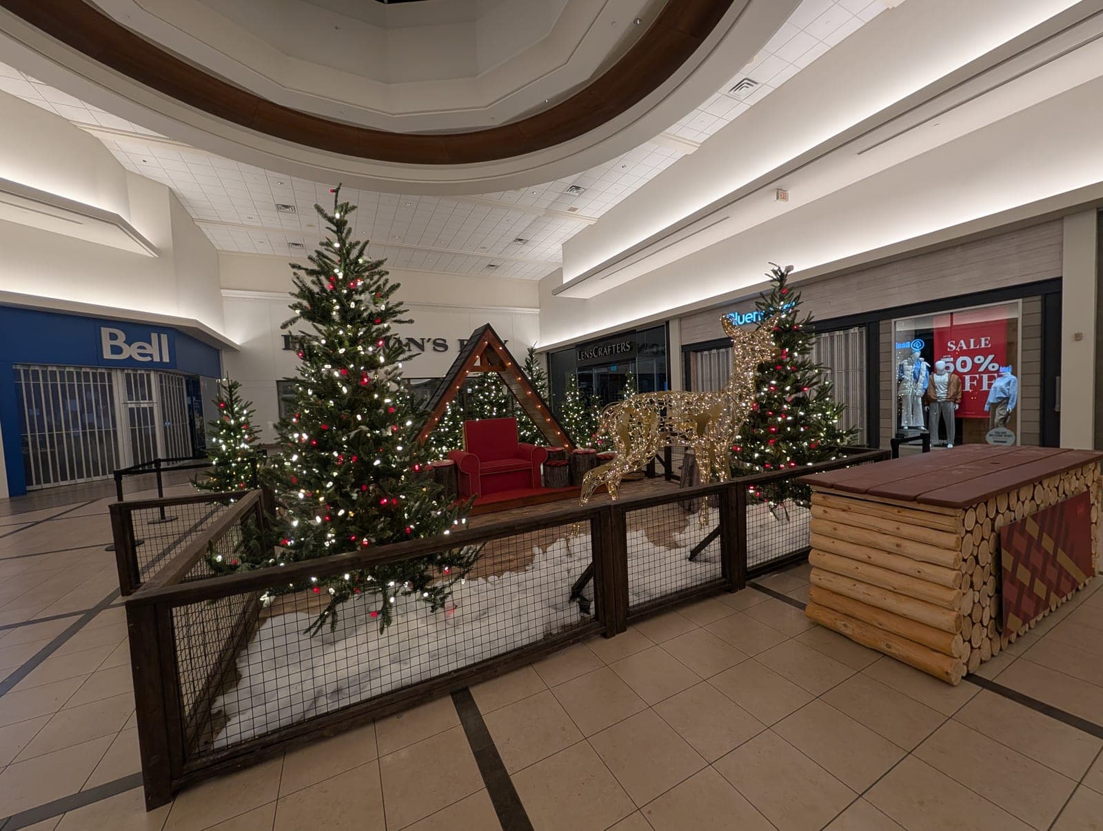 Rustic lumberjack-themed mall Christmas display with lit evergreen trees, a gold reindeer and an A-frame photo set