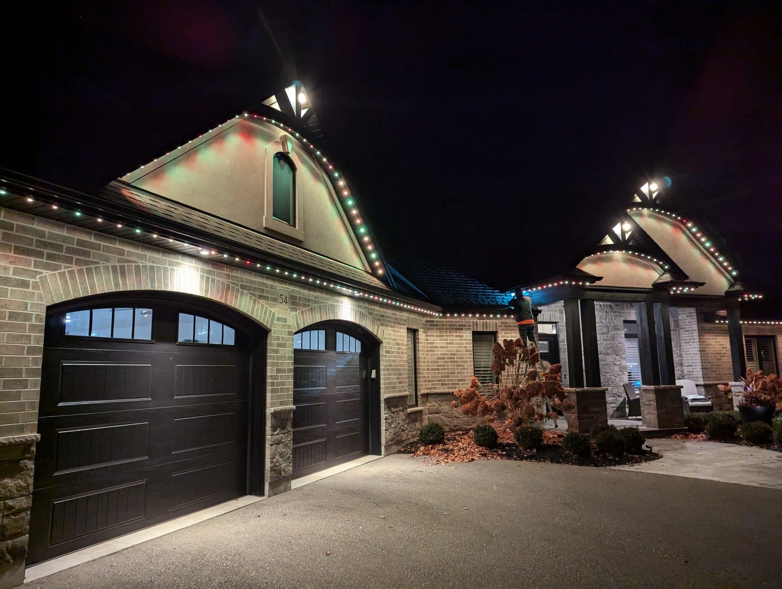 Multicolour C9 Christmas lights outlining the roofline and gable peaks of a two-tone brick home at night with an installer working near the front entrance.