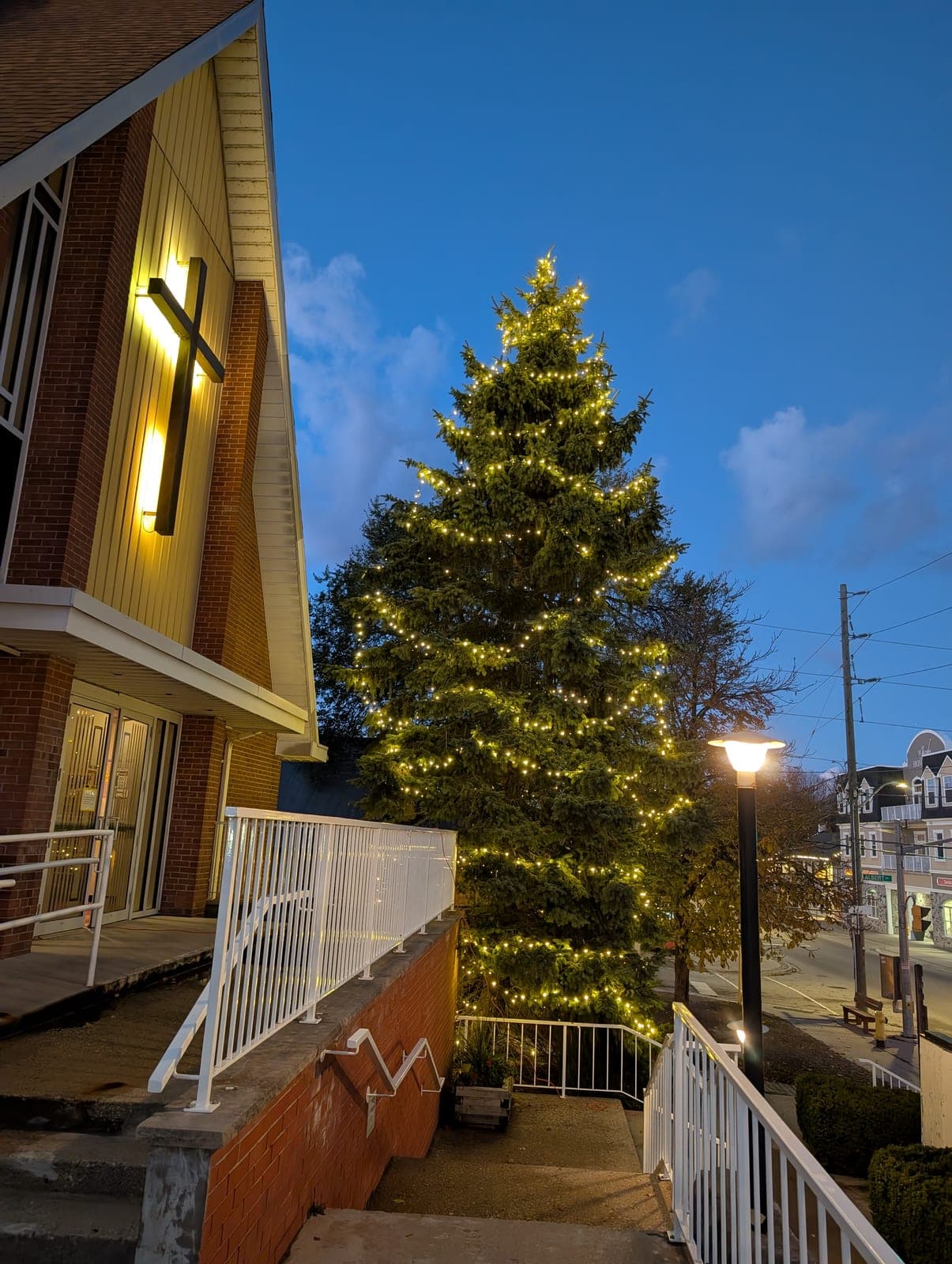 Tall evergreen tree wrapped in warm-white lights beside a church at dusk