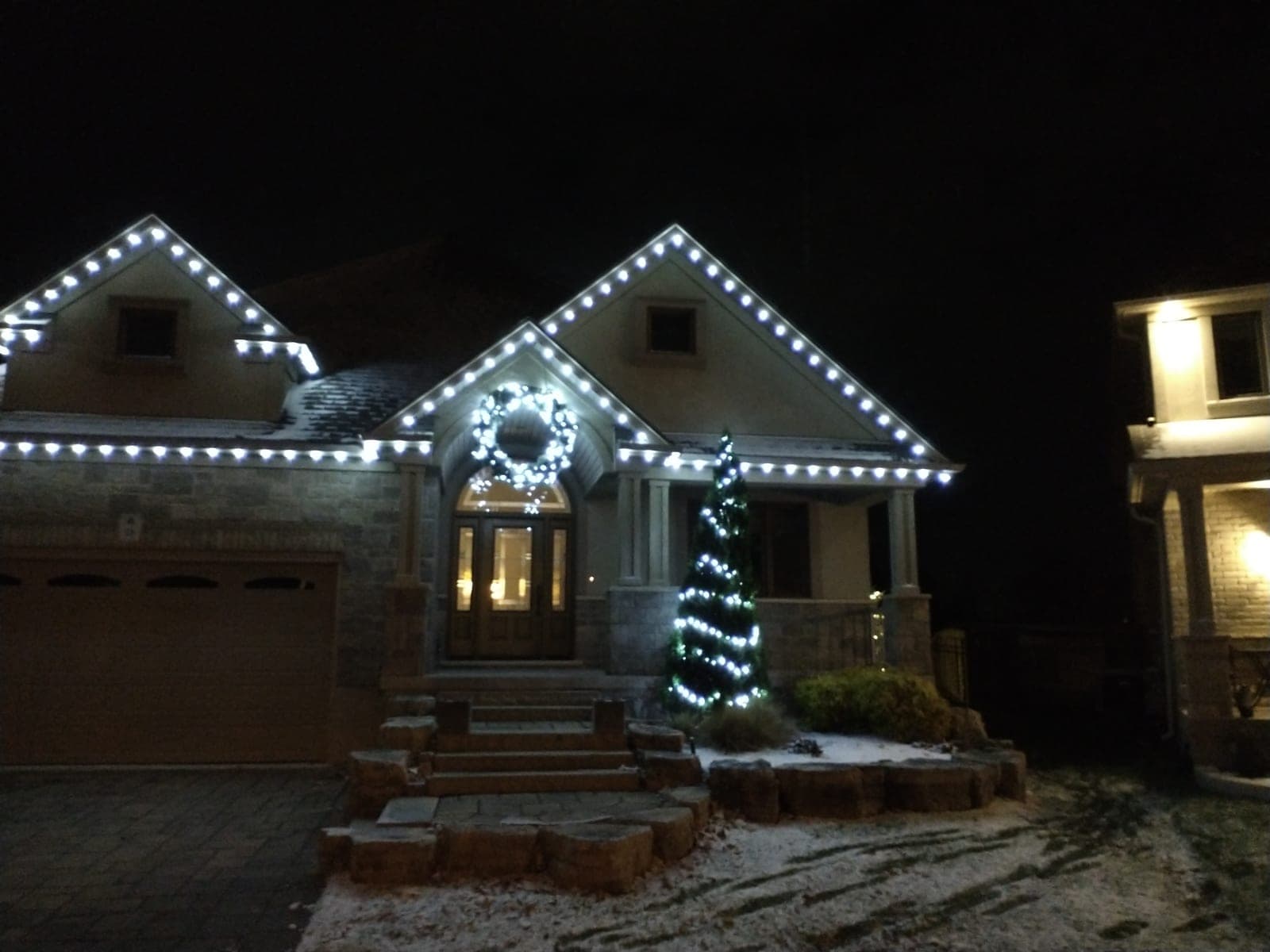 Stone bungalow with warm-white roofline Christmas lights, a lit wreath and a lit porch tree at night
