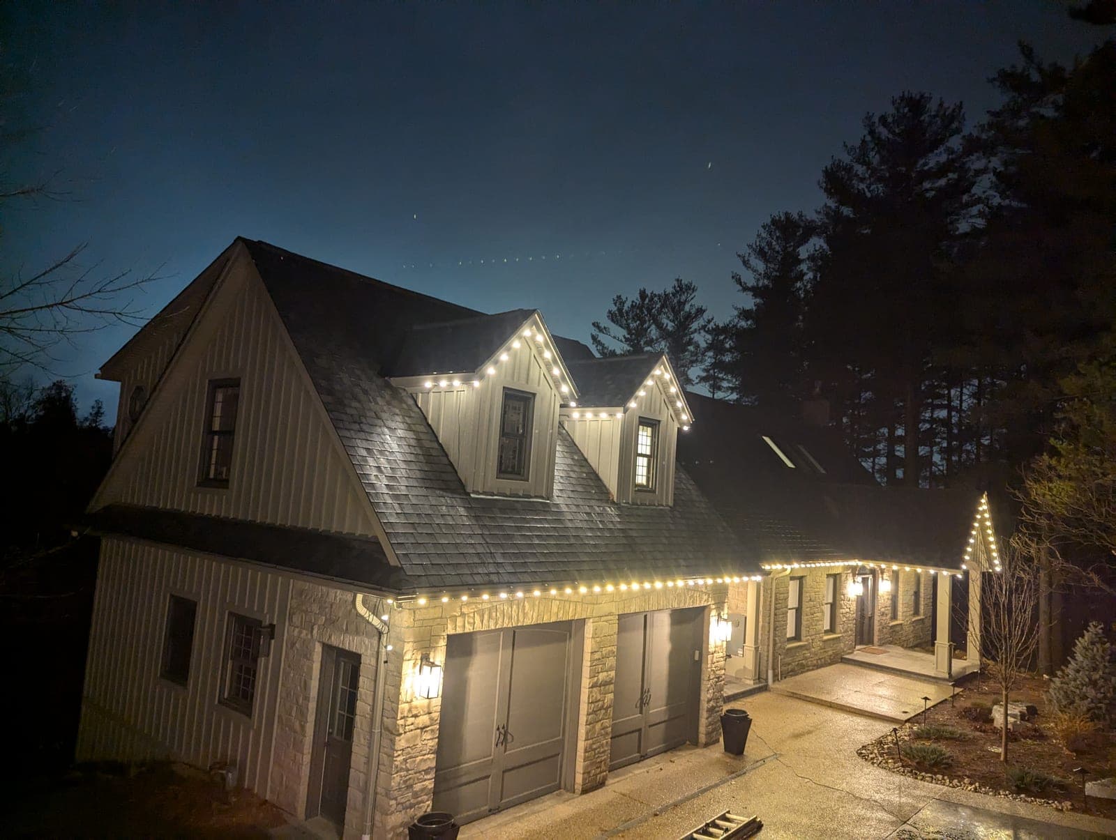 Two-storey stone and board-and-batten home outlined in warm-white roofline Christmas lights at dusk