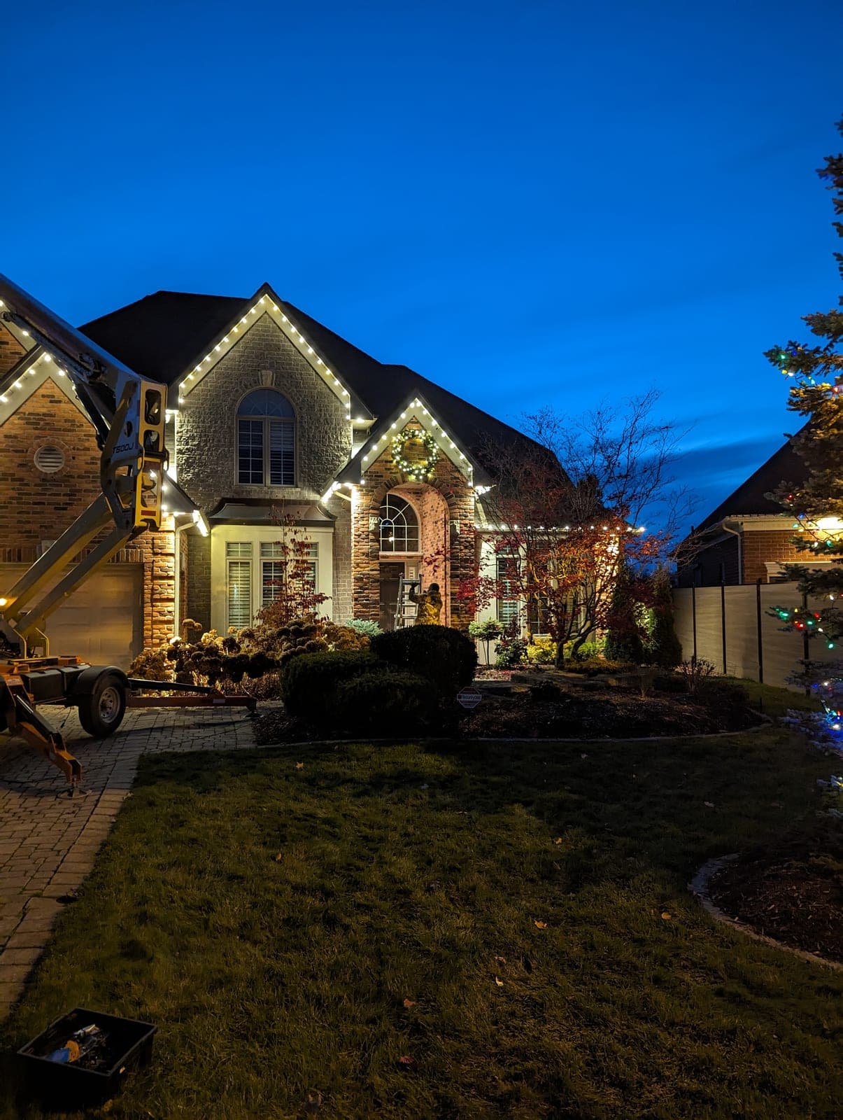 Two-storey stone home outlined with warm-white roofline Christmas lights and a lit wreath at dusk